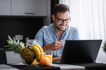 Handsome man using laptop computer at home, drinking juice and coffee in the morning. Working From Home.