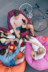 Sticker - high angle view of friends clinking with bottles of beer and holding fast food on bean bag chairs