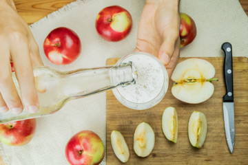 Pouring sparkling apple cidre drink into glass, top view. Flat lay image with hands fixing a drink of cider on rustic wooden table background with ripe apples