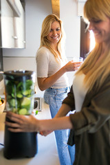 Wall Mural - Beautiful young woman preparing detox juice in the blender and talking with friend at home.