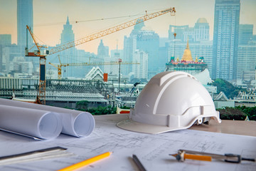 Architectural drawing plan with blueprint rolls, safety helmet and drawing tools on working table. Blurred crane and construction site in Bangkok in the background. Building construction concept