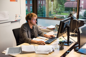 Young phd student studying writing final paper for exam at university. Working man at office desk typing at desktop computer in library room.