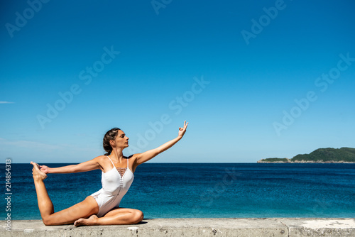 Fotografia Attractive woman in her 40s, practicing yoga on sea side, doing One Legged King Pigeon Pose - Eka Pada Rajakapotasana, in white swimsuit, with sea in the background