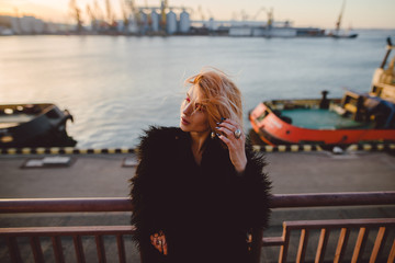 Young hipster woman with blonde hair, colorful make-up in grunge black coat standing on the bridge near the industrial port zone. Urban background. Fashion street style