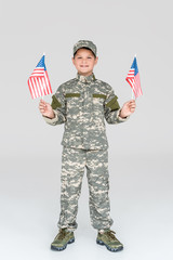 Wall Mural - smiling child in military uniform with american flagpoles in hands looking at camera on grey backdrop