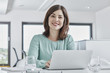 © Westend61 - Portrait of smiling young businesswoman using laptop at desk in office