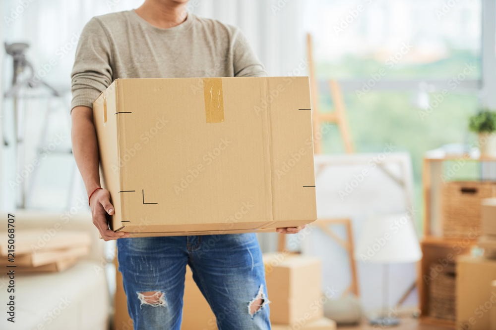 Crop view of strong male in casual clothes carrying large cardboard box on moving day with cartons and drawing easels on blurred background of art workshop. - obrazy, fototapety, plakaty 