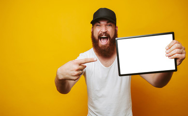 Cheerful amazed happy bearded hipster man in white t-shirt pointing at white screen on tablet at copyspace