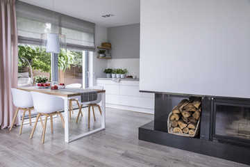 Modern fireplace and wood in a spacious dining room interior with white chairs by a wooden table and big windows