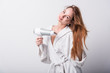 © Siarhei Kulikou - Beautiful red-haired girl in a white bathrobe bathing her hair with a hair dryer on a light background