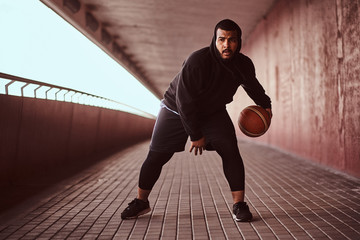 Wall Mural - Portrait of a dark-skinned guy dressed in a black hoodie and sports shorts playing basketball on a footway under bridge.