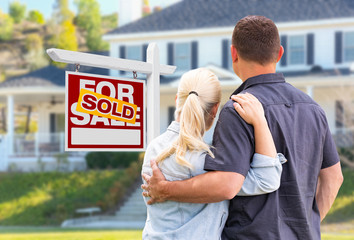 Young Adult Couple Facing Front of Sold Real Estate Sign and House