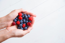 Fresh Blueberries And Raspberries In Women's Hands On A White Wooden Background.