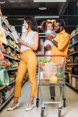 smiling african american man holding daughter while his wife choosing food with shopping list in supermarket