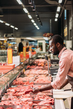 Focused African American Male Butcher In Apron Taking Steak Of Raw Meat In Supermarket