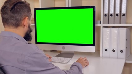Leinwandbilder - man sitting at workplace paying game on computer in small office. businessman with beard looking on display with green screen. employee in startup company typing on keyboard use pc mouse