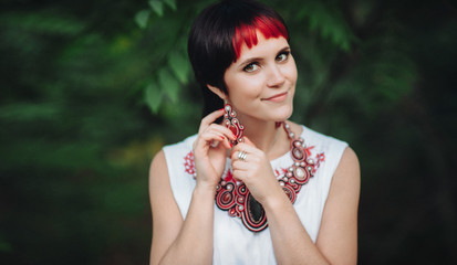 A beautiful girl on a green natural background adjusts her earring with her hand.The Ukrainian bride is dressed in ethnic style.Handmade jewelry.Original set of earrings and a necklace for the holiday