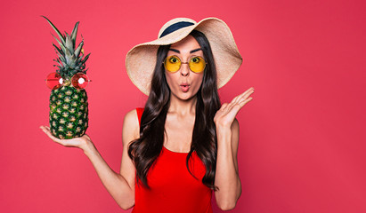 Poster - Happy young beautiful woman in red swimsuit, sunglasses and big summer hat with pineapple in sunglasses in hand posing over pink background