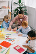 © LIGHTFIELD STUDIOS - teacher in eyeglasses helping multicultural preschoolers with drawing at table in classroom