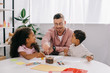 © LIGHTFIELD STUDIOS - smiling caucasian teacher in eyeglasses and african american kids drawing pictures with paints together in classroom