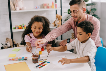 smiling caucasian teacher in eyeglasses and african american kids drawing pictures with paints together in classroom