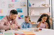 © LIGHTFIELD STUDIOS - teacher in eyeglasses and multiracial children drawing pictures with paints at table in classroom