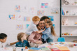 © LIGHTFIELD STUDIOS - interracial kids hugging happy teacher at table in classroom