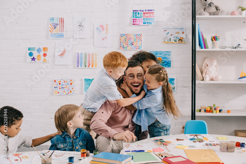 Fotografía interracial kids hugging happy teacher at table in classroom