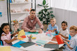 © LIGHTFIELD STUDIOS - male teacher and multiracial preschoolers sitting at table with colorful papers in classroom