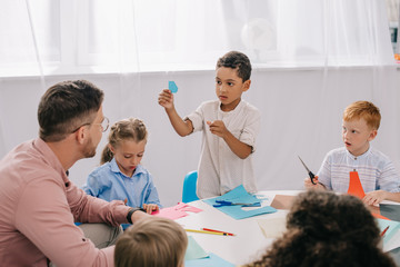 partial view of male teacher and multiracial preschoolers sitting at table with colorful papers in classroom