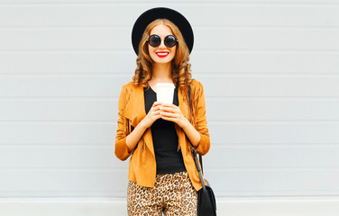 Elegant woman with coffee cup wearing retro elegant hat, sunglasses, brown jacket and black handbag over grey background