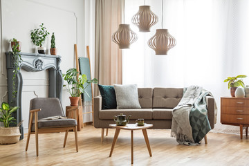 Real photo of a sofa with pillows and blanket standing next to an armchair and a small table in spacious botanic living room interior with wooden cupboard, shelf and plants