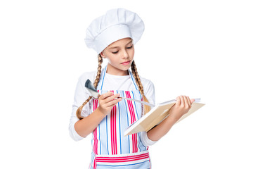 Wall Mural - schoolchild in costume of chef reading recipe book isolated on white