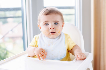 Portrait of cute adorable Caucasian child boy with dirty messy face sitting in high chair eating apple puree with fingers. Everyday home childhood lifestyle. Infant trying supplementary baby food