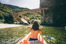 Paddler In A Canoe On A River In A Lush Green Valley Near A Medieval Village And Bridge
