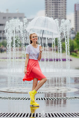 Young pretty girl with two braids in yellow boots and with transparent umbrella stands near fountain.