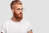Fototapeta  - Profile shot of brutal male with thick foxy beard, wears round glasses and looks thoughtfully aside, dressed casually, isolated over white background with blank space aside for your text or promotion