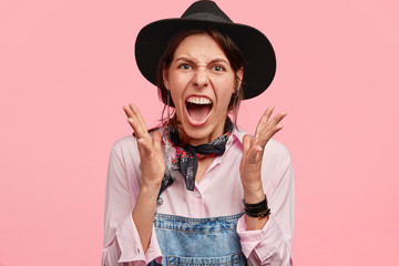 Wall Mural - Annoyed beautiful female farmer in black hat and denim overalls, gestures angrily and keeps mouth widely opened, has desperate expression, isolated over pink background. Negative facial expressions