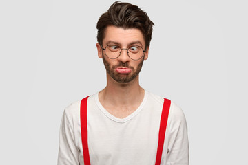 Wall Mural - Funny unshaven male crosses eyes and purses lips, makes grimace at camera, has comic expression, foolishes indoor against white studio background. Stylish bearded Caucasian man has nothning to do.