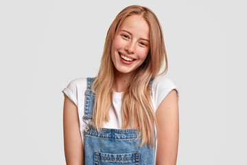Wall Mural - Beautiful happy Caucasian female with charming smile, dressed in fashionable overalls, has long light hair, expresses positive emotions as sees something pleasant in front, stands against white wall