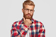 Attractive bearded man with ginger beard, holds chin, looks thoughtfully at camera, contemplates about something or how solve problem, dressed in checkered shirt, isolated over white background.