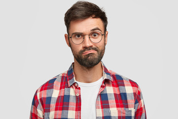 Headshot of puzzled hesitant displeased unshaven young guy purses lips, has uncertain exression, trendy haircut, wears checkered shirt, isolated over white background. Facial expressions concept