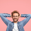 © wayhome.studio  - Vertical shot of relaxed carefree man keeps hands behind head, has positive expression, smiles pleasantly, listens something with interest, dressed in fashionable shirt, isolated over pink studio wall