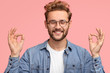 © WHstudio Leushin N - Portrait of cheerful satisfied male with stubble, stands in mudra sign, keeps eyes shut, has positive smile, stands indoor against pink background, wears jean shirt. People and body language concept