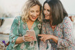 © Lomb - Two happy middle-aged women friends using a smartphone together outdoors. Concept of social media, digital connection, sharing memories, and a cheerful modern lifestyle for people in their 40s.