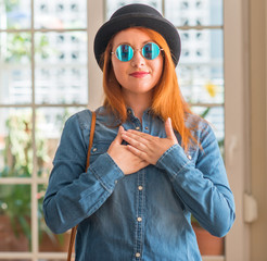 Sticker - Stylish redhead woman wearing bowler hat and sunglasses smiling with hands on chest with closed eyes and grateful gesture on face. Health concept.