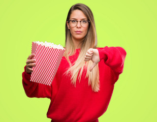 Wall Mural - Beautiful young woman eating popcorn with angry face, negative sign showing dislike with thumbs down, rejection concept