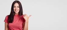 Optimistic Young Female With Toothy Friendly Smile Points Aside With Thumb, Shows Blank Space For Your Advertising Content Or Promotional Text, Has Long Dark Hair, Isolated Over White Background