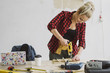 © Dash - Beautiful smiling young blond woman in casual clothes drilling small piece of wood on carpenter workbench with tools, instruments and laptop on background of white plastered wall