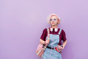 Portrait of a stylish bright young girl with headphones, sunglasses and colored hair on a purple background. Hipster girl on the background of a purple wall.